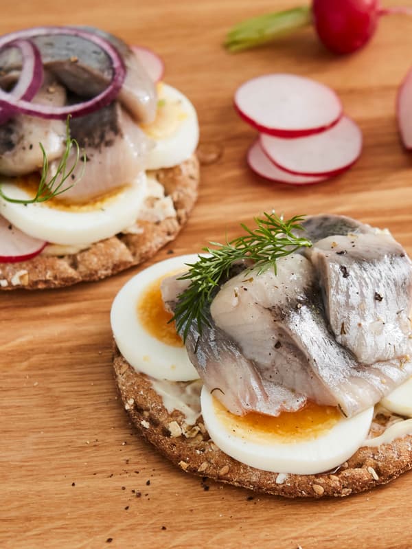 Dva kusy chrumkavého chleba KNÄCKEBRÖD FLERKORN obložené vajíčkami, marinovanými sleďmi SILL DILL a kôprom alebo cibuľou, v pozadí sú reďkovky.