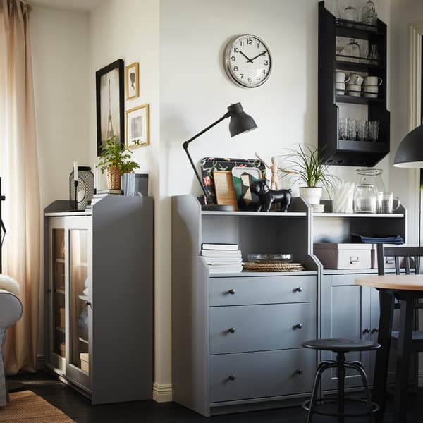 Dva siva fiokara s policama na kojima stoje ukrasni predmeti, zatim okrugli sto, crna stolica i crna visilica.