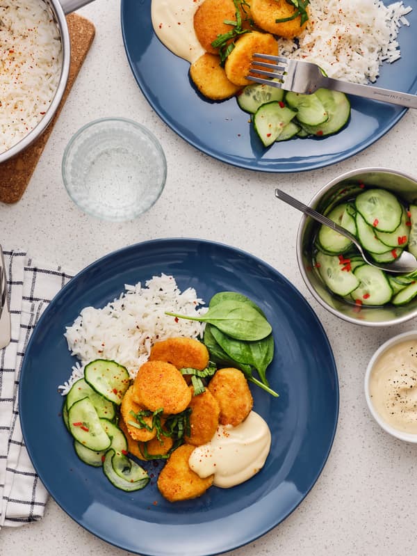 Dva tmavomodré taniere s pšeničnými nugetami, ryžou a uhorkovým šalátom.