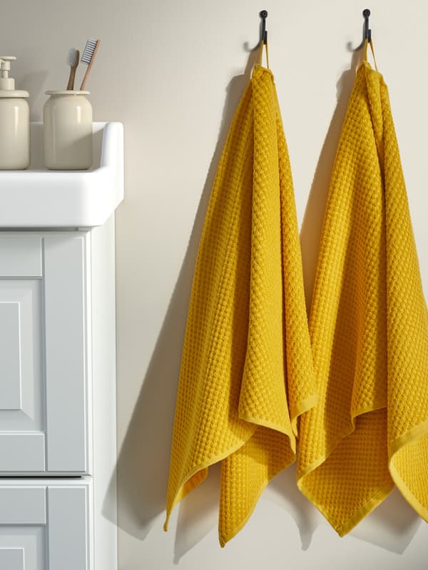 Dva žlté uteráky GULVIAL visia na stene vedľa bielej kúpeľňovej skrinky TÄNNFORSEN.
