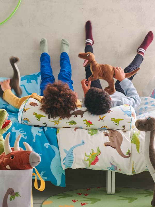 Dvě děti leží na posteli s povlečením JÄTTELIK, hlavy si opírají o polštář JÄTTELIK a hrají si s plyšovými dinosaury JÄTTELIK.