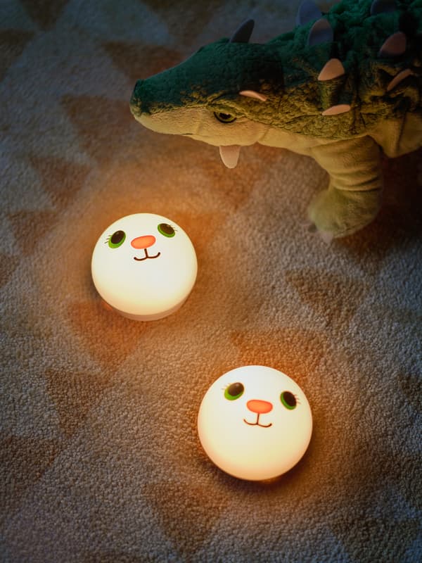 Dve nočné svietidlá KORNSNÖ na detskom koberci s pestrým vzorom a dinosaurom JÄTTELIK v pozadí.