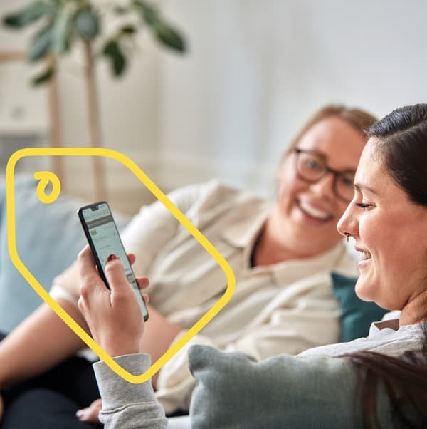 Dvě ženy sedící na pohovce a sméjící se uprostřed konverzace. Jedna z nich se dívá do telefonu, kolem kterého je namalovaný žlutý obrázek cenovky.