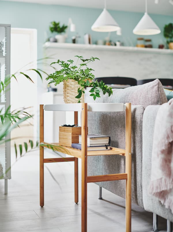 Dvije male biljke u teglama uz izrazito svijetlo rješenje za odlaganje na SATSUMAS stalku za biljke pokraj sofe u dnevnoj sobi.