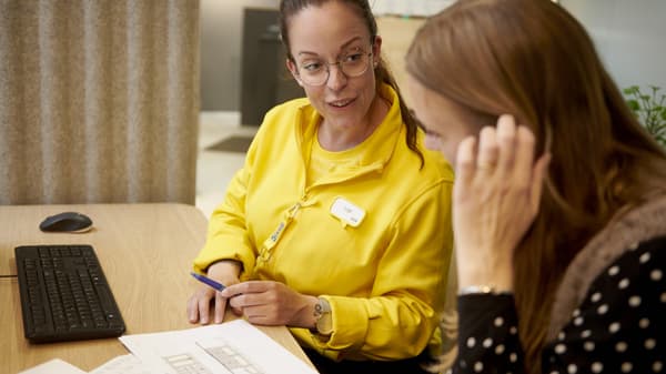 Dvoje ljudi sjedi za stolom, jedan u žutoj jakni pokazuje olovkom na papir s nacrtima, na stolu je tipkovnica.