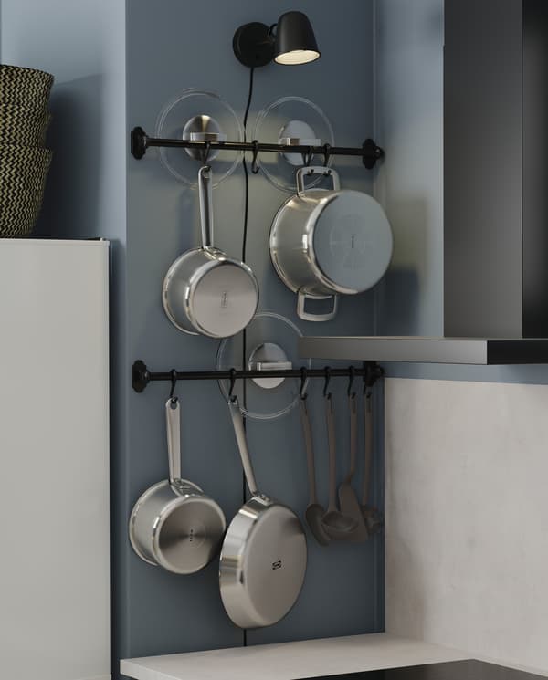 Dwa czarne drążki z haczykami, na których zawieszono rondle, garnki, patelnie i pokrywki. Nad drążkami wisi zapalona czarna lampa ścienna.