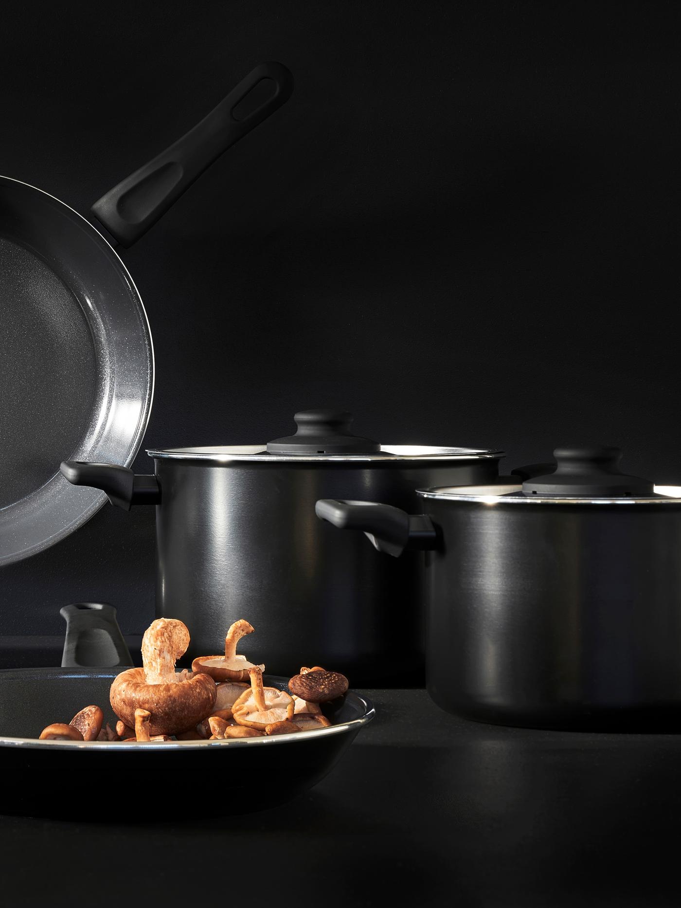 Dwa czarne garnki o różnych rozmiarach i patelnia HEMLAGAD stoją na czarnym bacie, z tylu jest czarne tło
