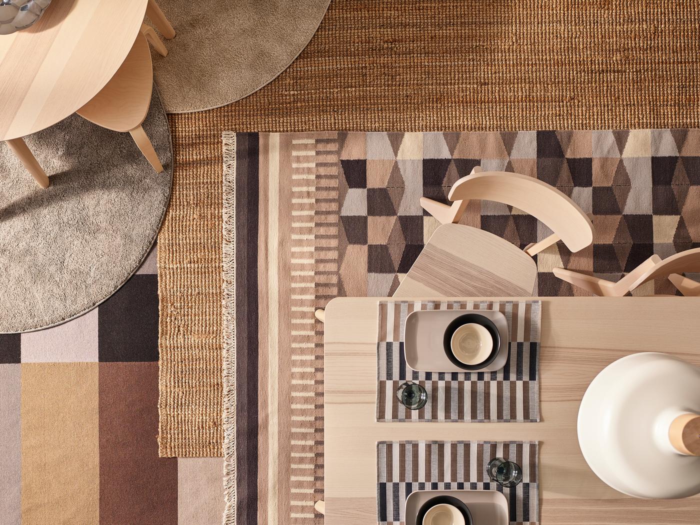 Dwa stoły z okleiną imitującą jesion i krzesła LISABO stoją na podłodze, na której leżą różne dywaniki. Wśród nich są dywanik LOHALS i kilka innych ozdobionych wzorami.