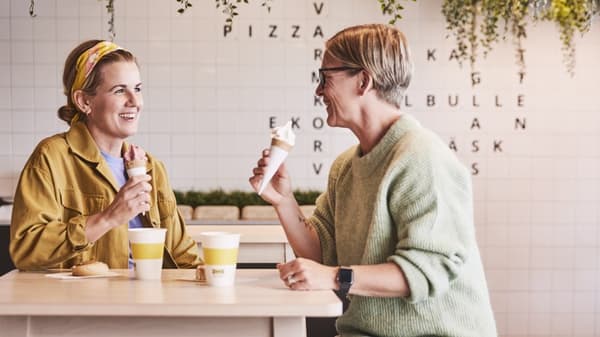 Dwie uśmiechnięte kobiety jedzą lody siedząc przy stole w bistro IKEA. Na stole znajdują się cynamonowe bułeczki oraz papierowe kubki z napojami. 