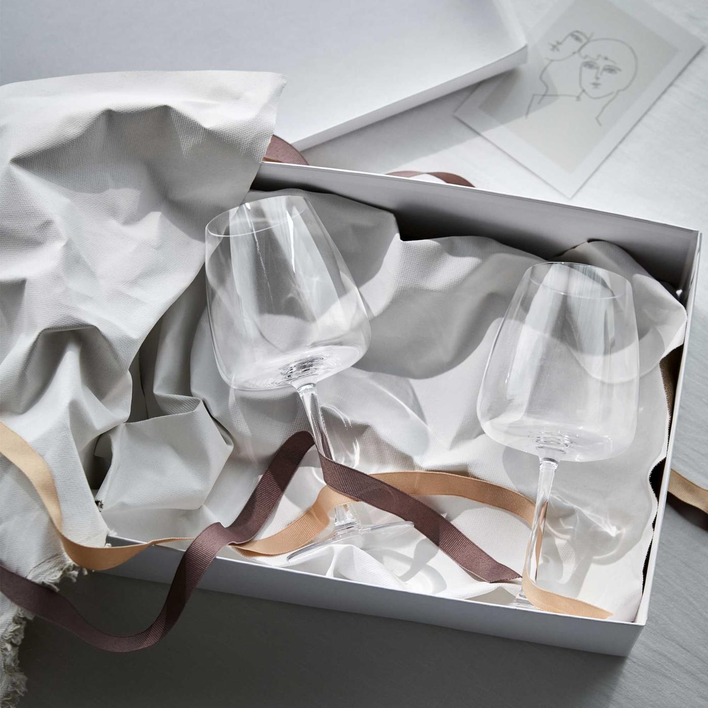 종이 포장지 여러 장이 담긴 종이 상자에 DYRGRIP 뒤르그리프 와인잔 두 개가 놓여 있고, 그 위에 놓인 컬러 끈이 우아한 분위기를 연출하는 모습.