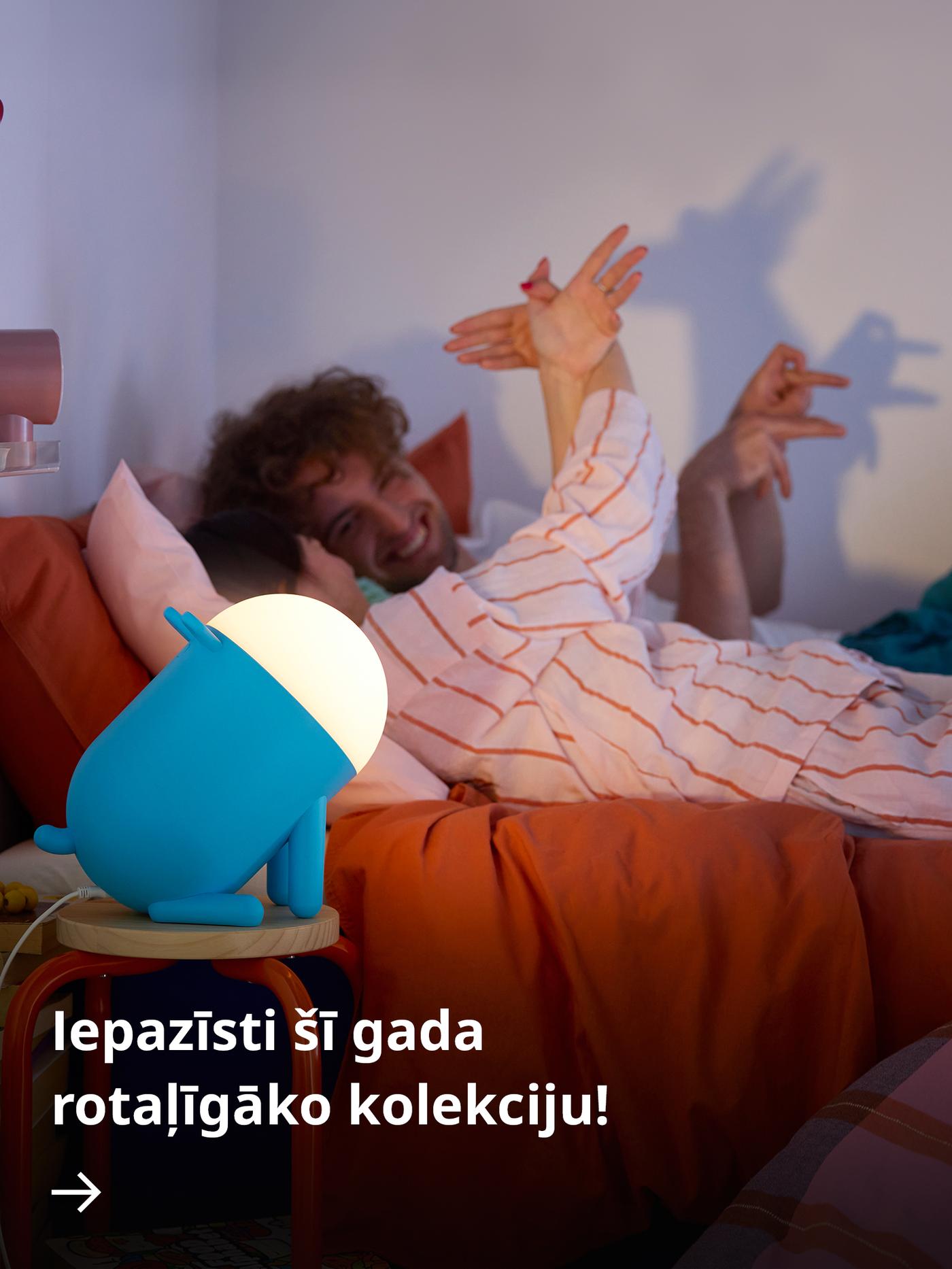 Dzeltena žirafes formas GREJSIMOJS grīdas lampa novietota tumšā istabā blakus bērna gultai.
