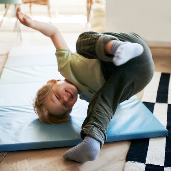 Dziecko w ciemnozielonych spodniach i jasnozielonej koszulce wykonuje przewrót przez głowę na niebieskiej składanej macie gimnastycznej PLUFSIG.