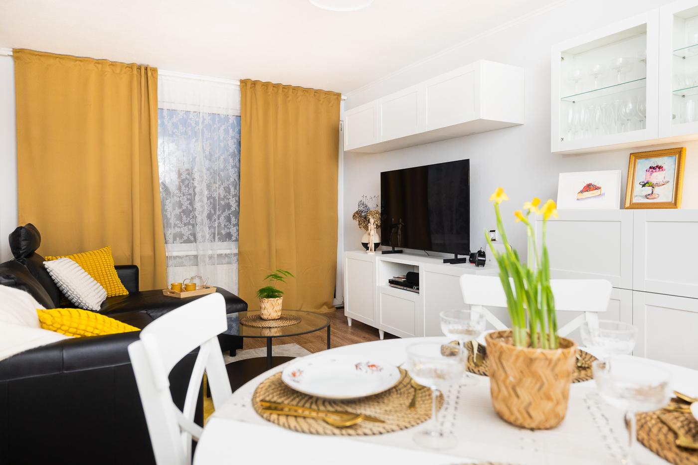 Dzīvojamā istaba pēc pārvērtībām ar gaišām mēbelēm, dzelteniem aizkariem un skaidri nodalītām zonām – redzama ēdamzona priekšplānā un atpūtas zona ar dīvānu un televizoru fonā. Telpa izskatās gaiša, sakārtota un mājīga.