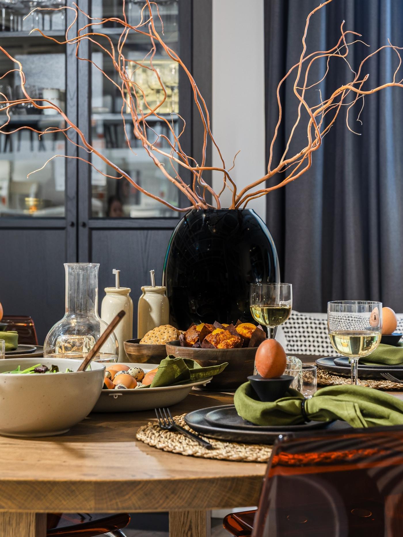 Easter table arrangement with earthy plates, green linen napkins and spring branches on a bare wooden table.