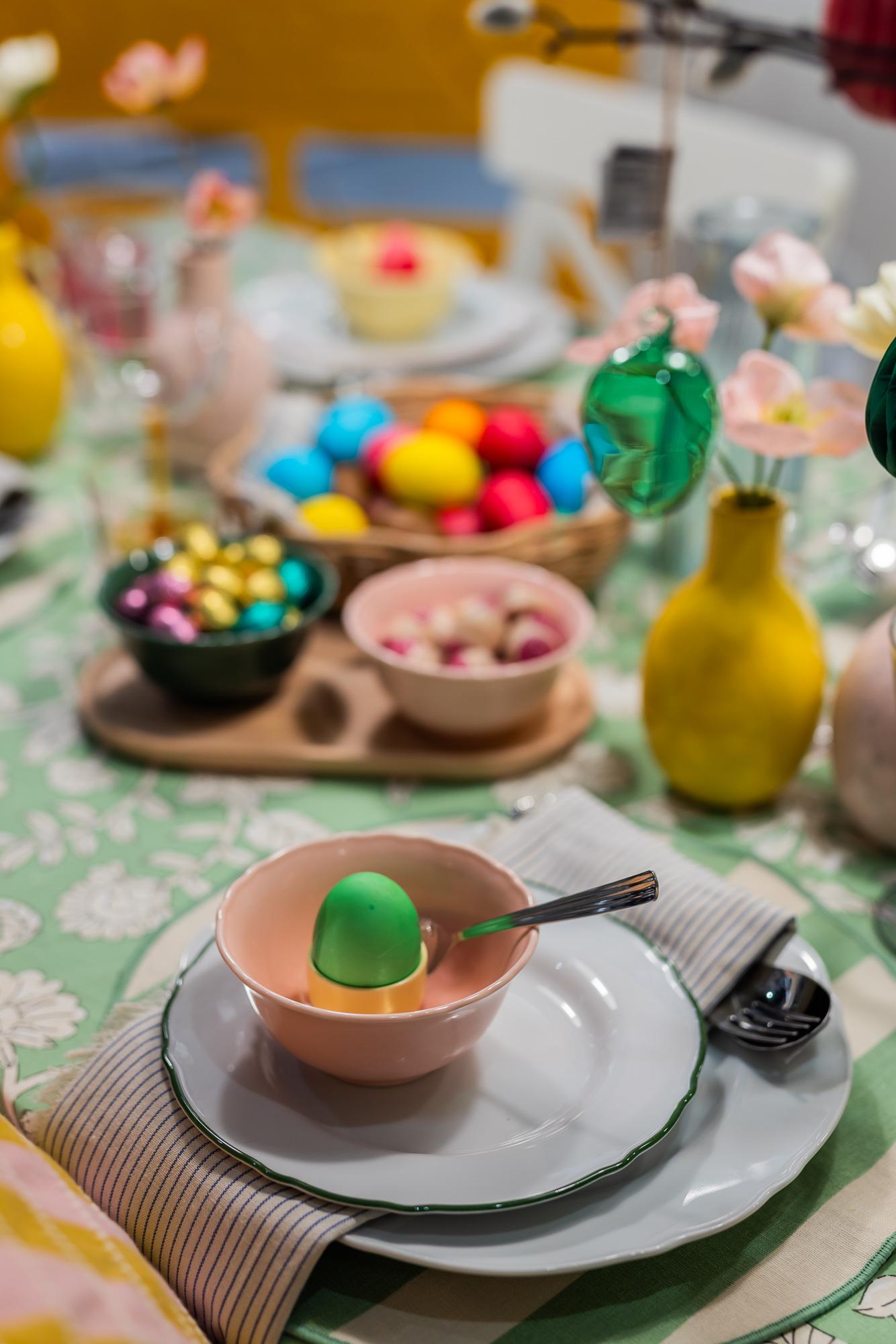 Easter table setting: white plates of various sizes stacked on one another topped with a small bowl with a green egg. In the background - an arrangement of Easter and chocolate eggs in small bowls on a wooden tray.