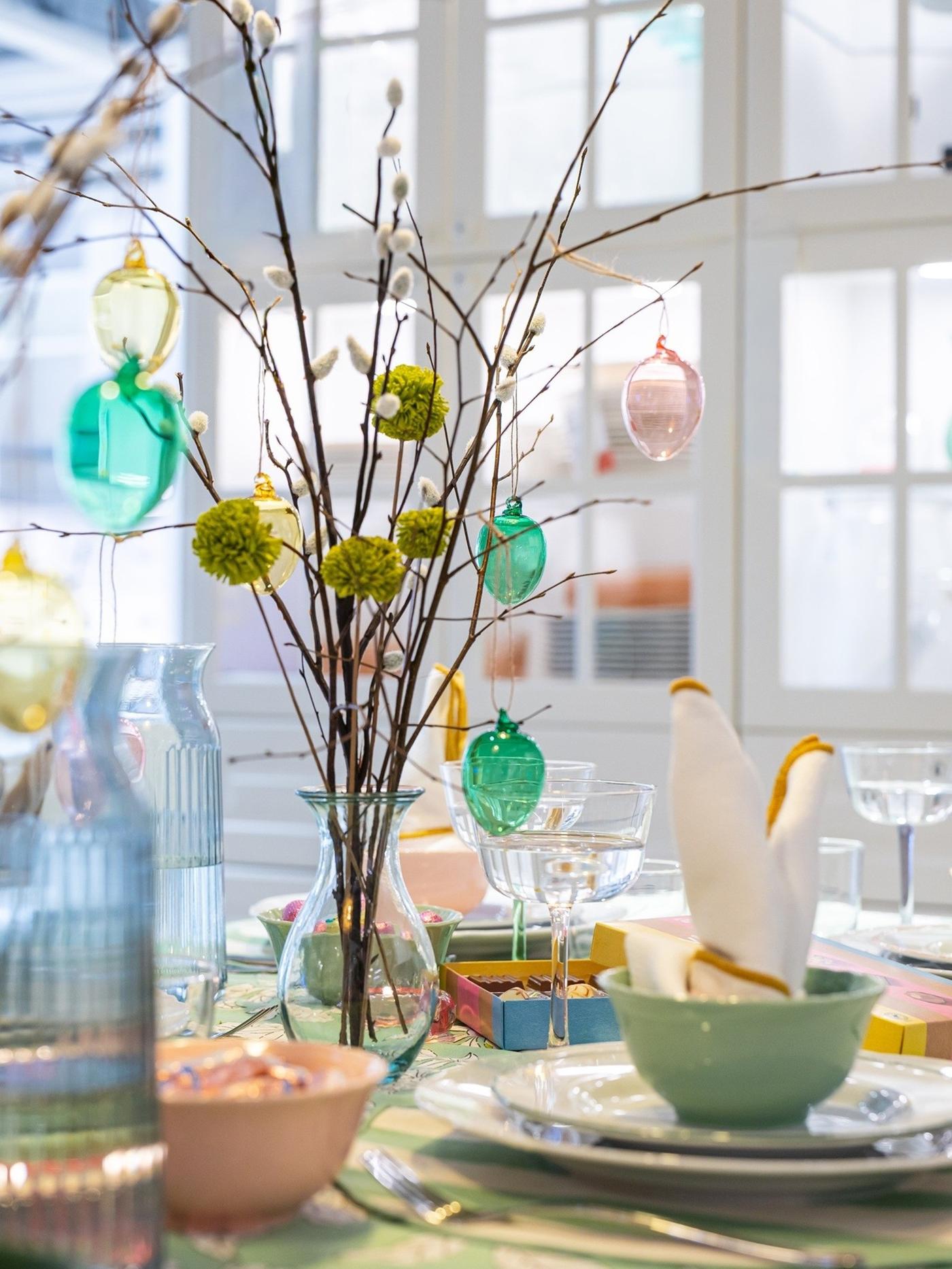 Easter table setting with white plates, green bowls and a floral tablecloth, decorated with budding branches and hanging pastel eggs.