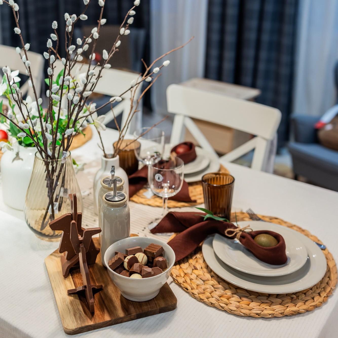 Easter table with a white tablecloth, a glass vase with willow branches, a wooden tray with spice jars, and chocolate treats, surrounded by white chairs.
