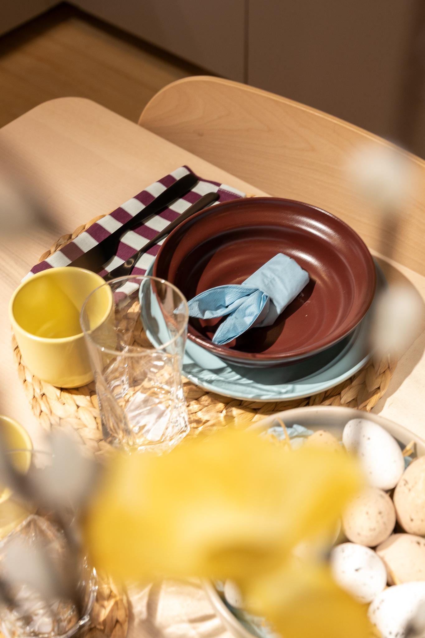 Easter-themed dining table with layered tableware, yellow mugs and floral arrangements arranged in glass vases, seen reflected in a large round mirror with a kitchen in the background.