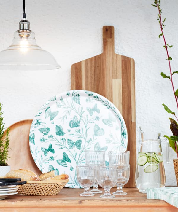 Ebédhez megterített asztal, poharakkal, kenyérrel, szalvétákkal, tányérokkal, zöld tálcával és fa vágódeszkával.