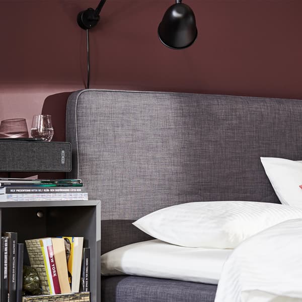 Een boxspring in donkergrijs, wit beddengoed, een lichtroze sprei en zwarte wandlampen.