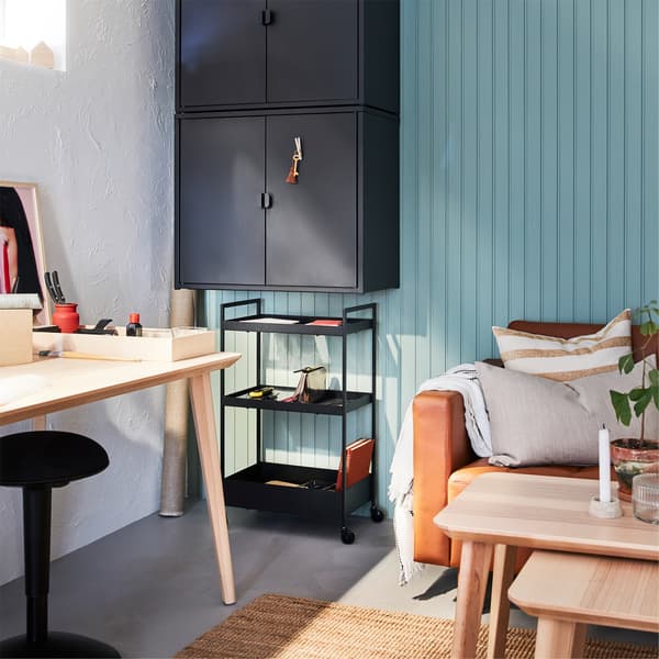 Een bureau in essenfineer, een jute vloerkleed, zwarte wandkasten, een zwarte trolley en een goudbruine leren bank.