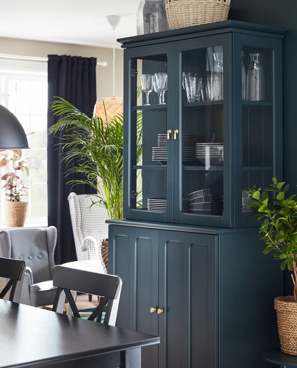 Een donker blauwgroene LOMMARP kast met vitrinedeuren staat naast een eettafel met eetgerei achter de vitrinedeuren.