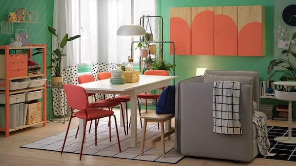 Een eetkamer met een witte tafel, rode stoelen, een grijze bank, een kast en een koraalrood geverfde stellingkast.