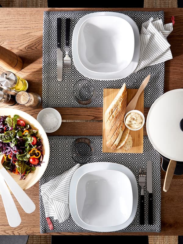 Een eettafel met witte borden en kommen, placemats met patroon, bestek, glazen en een saladekom.