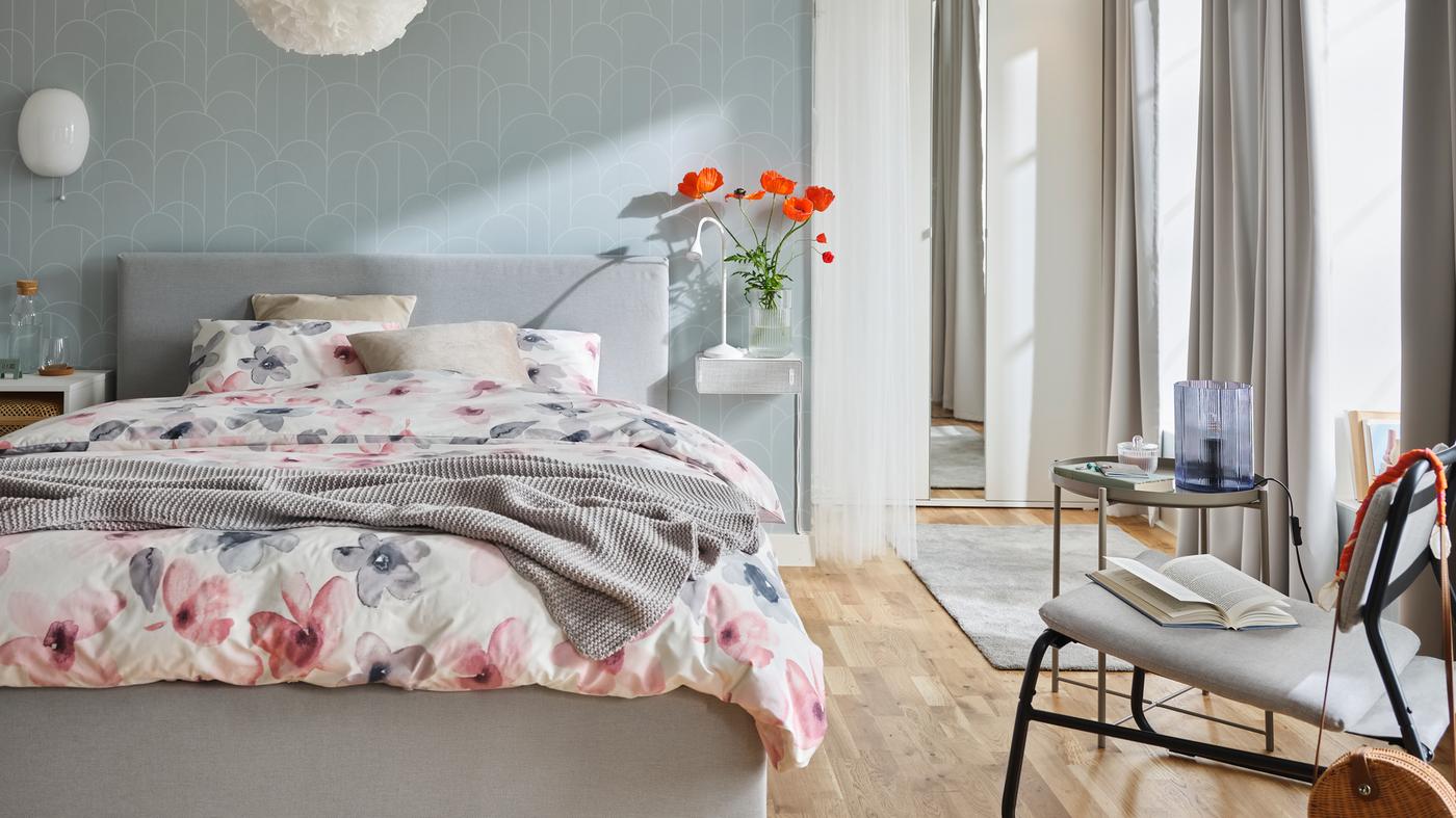 Een GLADSTAD bed in een grijze slaapkamer, gedekt met LAPPNYCKLAR beddengoed met een bloemenpatroon. Naast het bed staan een fauteuil en een tafel.