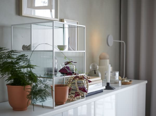 Een glazen IKEA SAMMANHANG vitrinekistje met witte kaders en plankjes op een buffetkast met planten, potten, boeken en decoratiestukken.