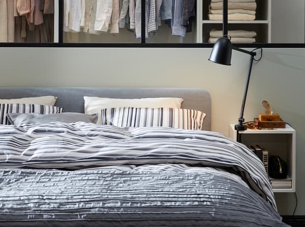 Een grijs, gestoffeerd bedframe, gestreept beddengoed, een wit nachtkastje en zwarte lampen