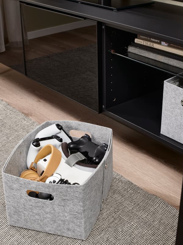 Een grijze opbergdoos van vilt gevuld met verschillende gamecontrollers, geplaatst op een grijs vloerkleed voor een zwart tv-meubel.