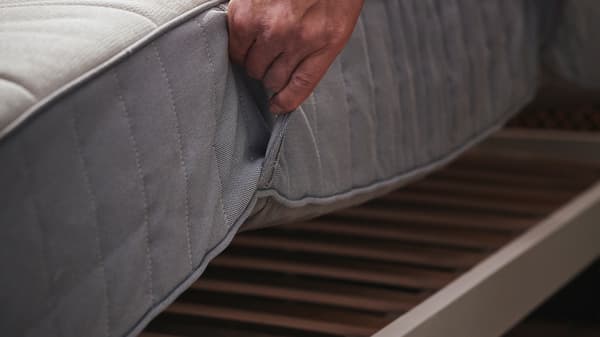 Een hand die een stevige/lichtblauwe VALEVÅG pocketveringmatras van een bedframe tilt.