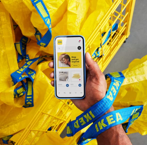 Een hand houdt een smartphone vast waarop de IKEA app zichtbaar is. Rondom liggen gele IKEA-tassen met blauwe handvatten en een gele winkelwagen op de achtergrond.