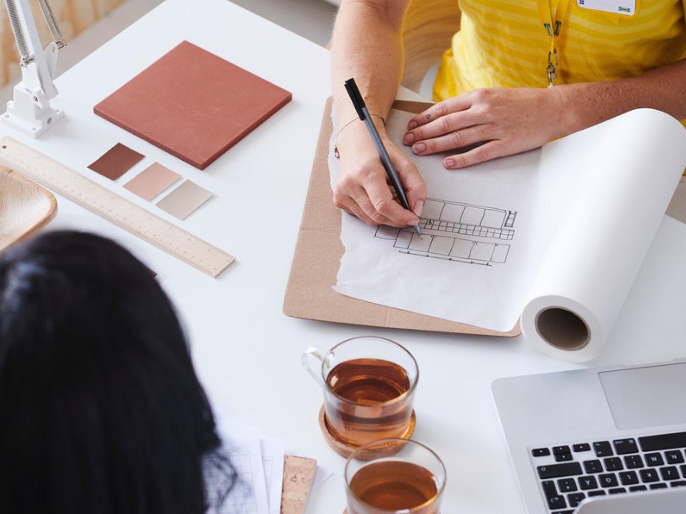 Een IKEA interieur designer heeft een interview met een klant aan een wit bureau met allerlei kleurstalen, schetsen, glazen theekopjes en een witte bureaulamp.