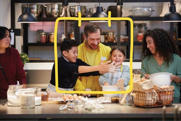 Een IKEA medewerker bij een aanrecht met kommen, eieren en bloem lacht, twee kinderen en hun moeders zijn aan het bakken.