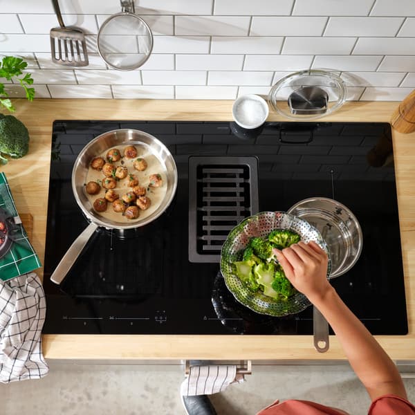 Een inductiekookplaat met geïntegreerde afzuigkap. Hier worden broccoli en gehaktballetjes gekookt, en je ziet een hand van iemand boven de broccoli.