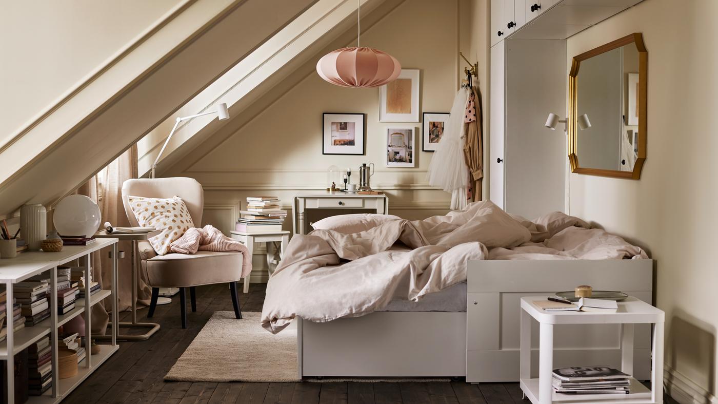 Een kamer met een verlengd wit daybed met lichtgrijs/beige bedlinnen, diverse opbergmogelijkheden, een beige fauteuil en roze hanglamp.