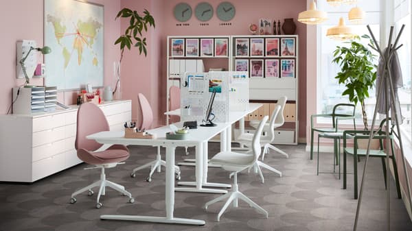 Een kantoorruimte met roze muren en witte BEKANT zit/sta-bureaus met HATTEFJÄLL draaistoelen.