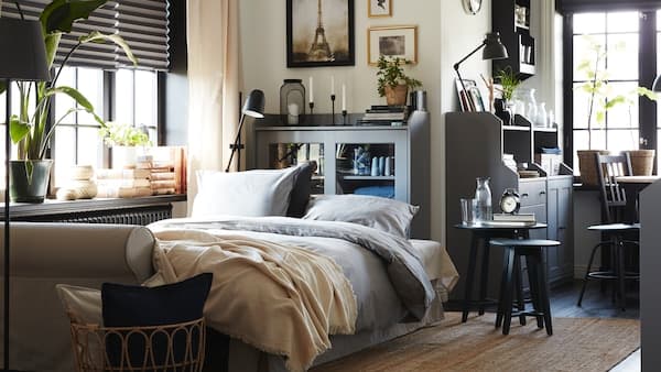 Een lichtbeige slaapbank opgemaakt als bed met grijs beddengoed. Daarachter staat een grijze kast met vitrinedeuren en een juten vloerkleed op de grond.
