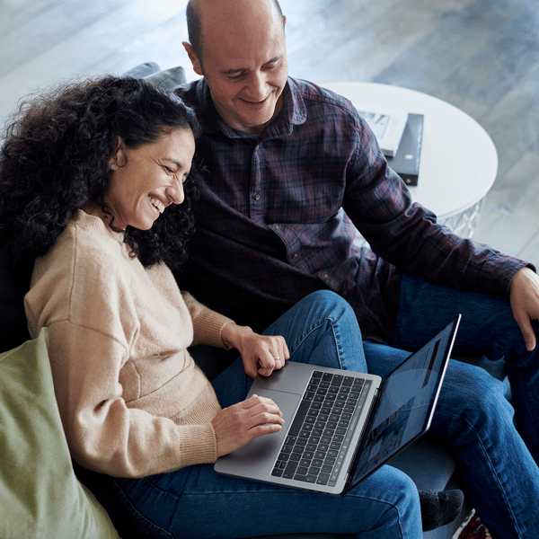 Een man en vrouw zitten lachend samen op de bank en kijken naar een laptop op de schoot van de vrouw.