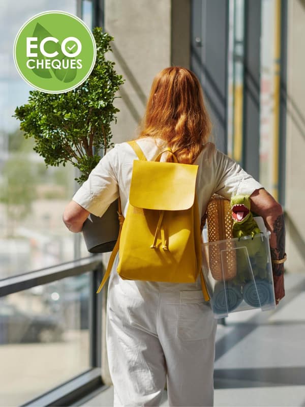 Een persoon met lang haar, gekleed in het wit en met een gele rugzak, door een gang terwijl die een kamerplant en een doorzichtige bak met boodschappen draagt, met linksboven een groen logo met de tekst "ECO CHEQUES".