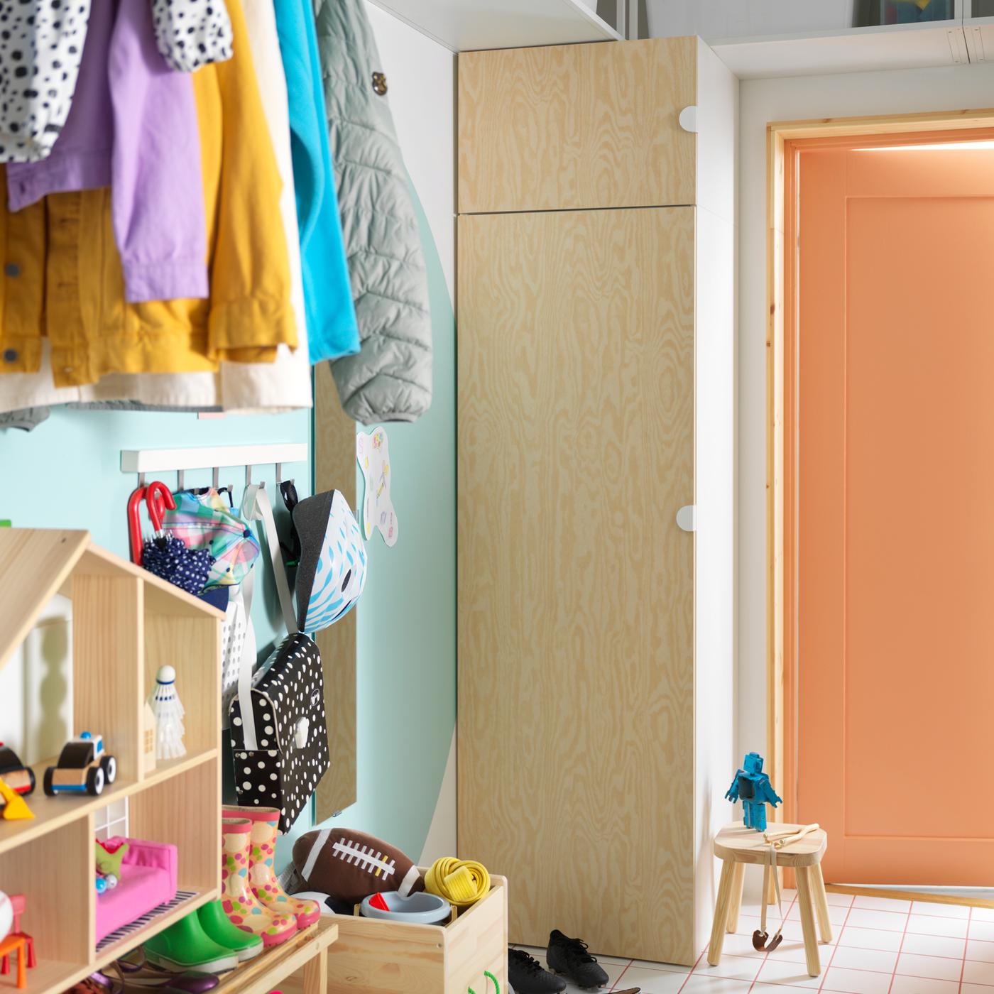 Een PLATSA garderobekast met KALBÅDEN deuren staat in een kleurrijke hal met een rij kinderschoenen, en veel kleren en speelgoed.