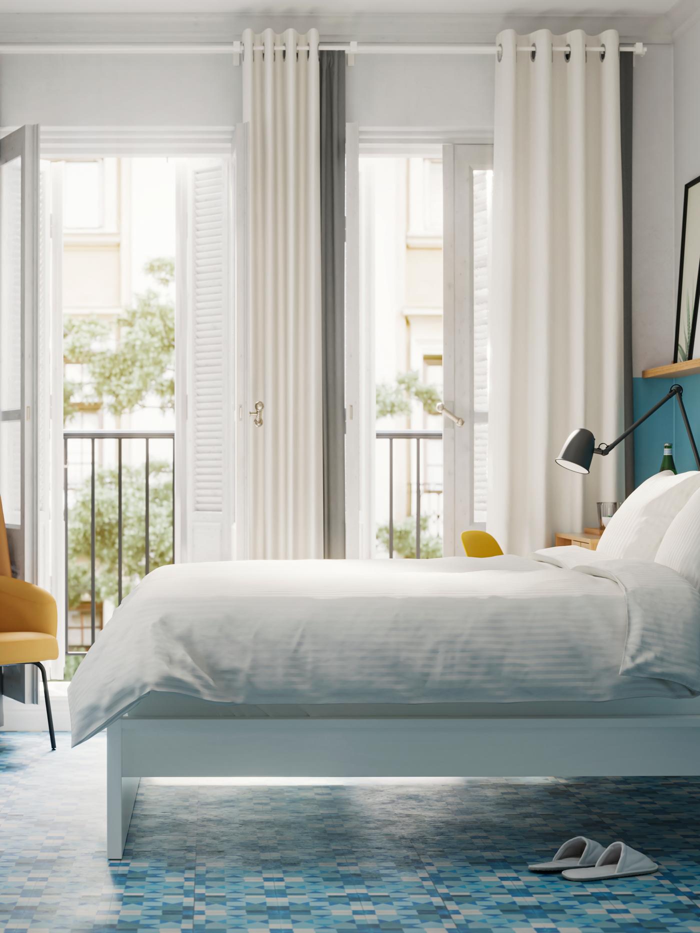 Een slaapkamer met een wit bed met wit beddengoed, blauwe muren en vloeren en openslaande deuren naar een balkon.