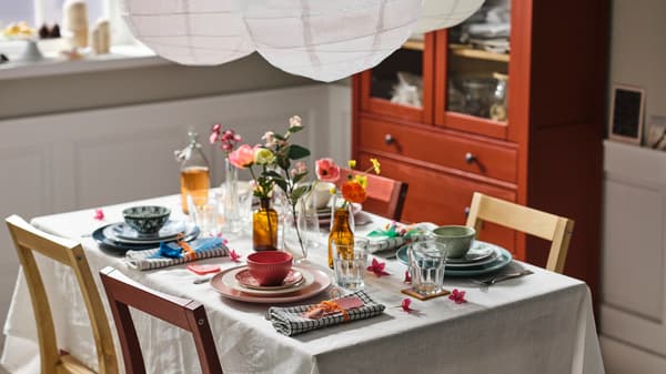 Een tafel met een naturel/beige tafelkleed gedekt met gemengd servies in verschillende kleuren, POKAL glazen en kunstbloemen...