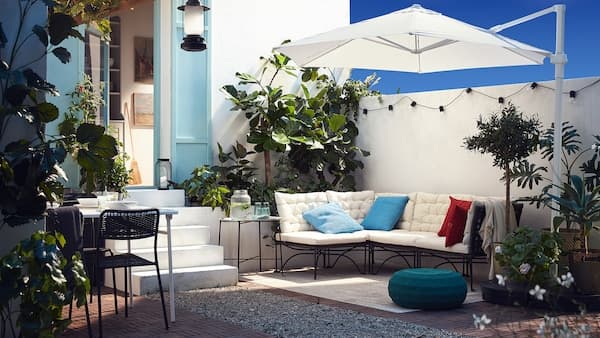 Een terras met een modulaire zitbank met witte kussens, een witte parasol, een blauwgroene poef en heel veel planten.