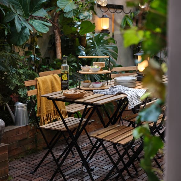 Een terras met veel groene planten, tuinstoelen en -tafels in acacia/zwart, een gele plaid en een dienblad in bamboe.