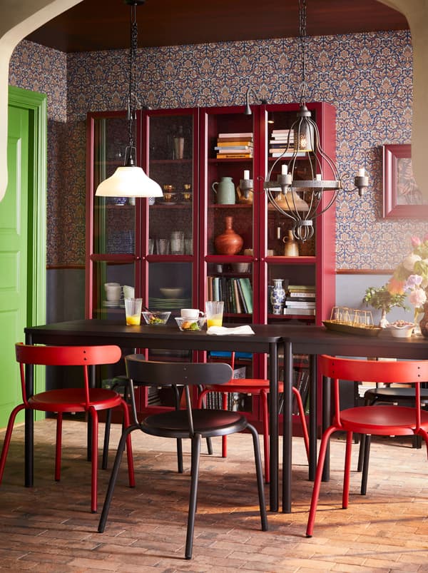 Een TOMMARYD eettafel is gecombineerd met vier kleuren YNGVAR stoelen. Erachter staan twee rode vitrinekasten.