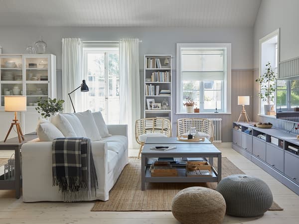 Een traditionele woonkamer met een witte bank, een jutetapijt, twee grijze salontafels, een grijze tv-bank en twee rotanfauteuils.