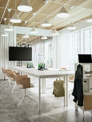 Een vergaderruimte met witte tafels, houten bureaustoelen met wielen, een grote flatscreen en een houten plafond met meerdere witte hanglampen. 